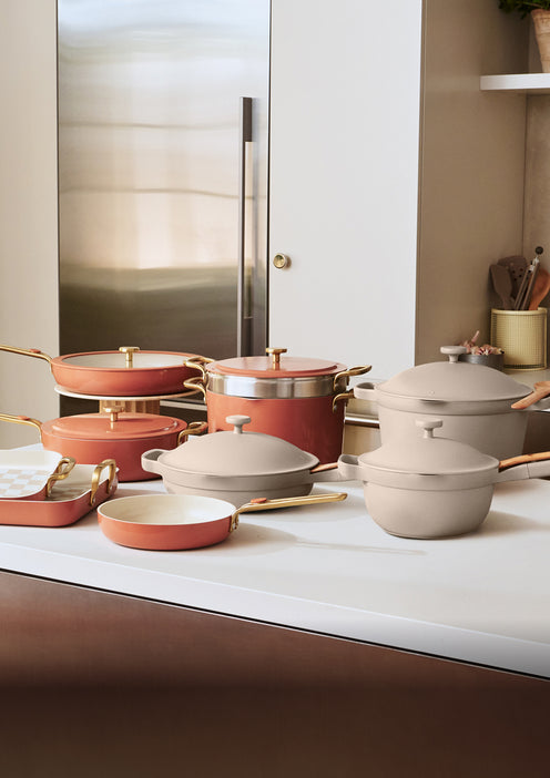 Assorted ceramic cookware in pink and beige with gold handles arranged on a white kitchen counter in front of a stainless steel refrigerator.
