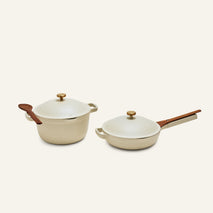 The Cookware Duo features two cream-colored pieces: a pot with lid and spoon, and a deep pan with lid and spatula, both with wooden handles, all set against a plain background.
