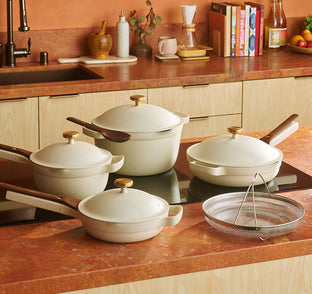 A set of cream-colored cookware with wooden handles is displayed on a kitchen countertop, including pans, pots with lids, and a metal strainer basket.