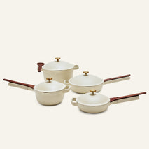 The Cookware Set includes four cream-colored pieces, featuring the Perfect Pot and Always Pan with wooden handles and gold knobs, elegantly displayed on a light background.