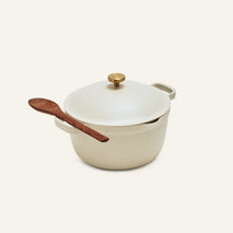 The Perfect Pot is an 8-in-1 cream-colored nonstick pot with a lid and gold knob, featuring a wooden spoon on one handle, set against a plain light background.
